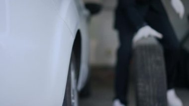 Close-up white car with blurred unrecognizable man in suit rolling tires from background indoors. Caucasian elegant driver changing tyres of vehicle in garage