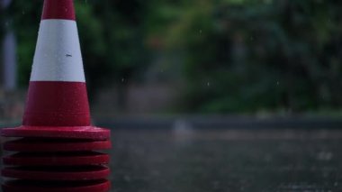 Red and white road cone on the left with rain falling on asphalt road at background. Rainy summer spring day outdoors with no people