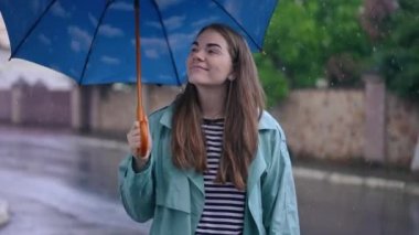 Joyful young woman standing in rain putting down umbrella smiling. Portrait of confident cheerful Caucasian lady posing and looking around on city street on rainy day