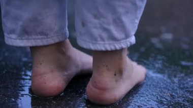 Close-up feet of barefoot young woman standing on asphalt road start walking on puddles outdoors in slow motion. Back view Caucasian lady strolling outdoors in rain