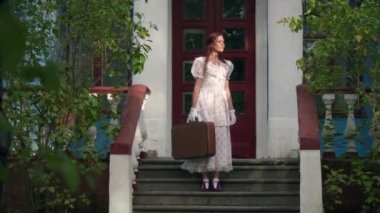 Wide shot beautiful young woman in retro dress with vintage suitcase standing on porch outdoors. Portrait of confident gorgeous Caucasian lady with luggage looking away smiling