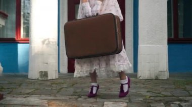 Unrecognizable woman in elegant retro dress and shoes standing with suitcase on porch. Young slim Caucasian vintage lady outdoors with baggage