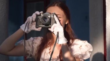 Close-up portrait of smiling talented retro woman taking photos on camera standing at golden sunset outdoors. Happy confident Caucasian lady in vintage dress and gloves enjoying hobby on porch