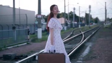 Tracking side view of elegant young lady crossing railroad lines looking around. Beautiful confident Caucasian time traveler with vintage suitcase in retro dress outdoors