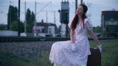 Wide shot beautiful lady in elegant retro dress sitting on travel bag looking away. Portrait of charming young Caucasian woman in vintage outfit on railroad outdoors