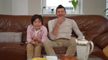 Beyaz çocuk ve baba TV 'de spor maçı izlerken hayal kırıklığına uğramış yüz ifadeleri sergiliyorlar. Oturma odasında kanepede oturan memnuniyetsiz bir adam ve çocuğun ön manzara portresi.