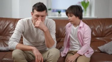 Orta ölçekli, düşünceli, beyaz bir adamın çocuk gibi kanepeye oturup el sıkışırken görüntüsü. Mutlu baba oğlunu evde karşılıyor.