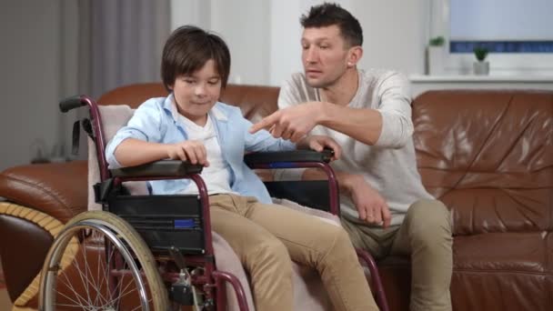 Tekerlekli Sandalyedeki Meraklı Çocuk Oturma Odasında Kanepede Oturan Adamla Konuşuyor — Stok video