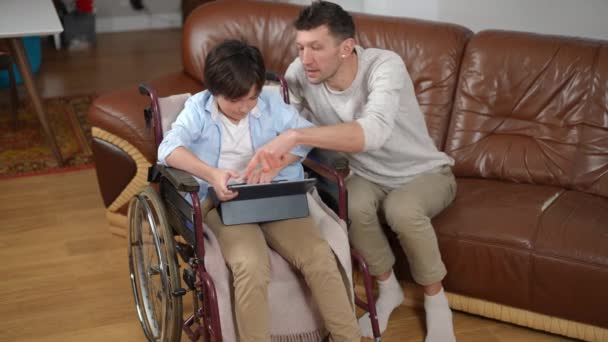 車椅子の息子と父親は デジタルタブレット上でサーフィンインターネットを話して吸収 高い角度ビューの広いショットの白人少年と男楽しみレジャースクロールソーシャルメディアゴシップ — ストック動画