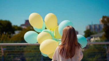 Ukrayna 'da cam köprünün üzerinde duran mavi ve sarı balonlu nakışlı esmer kız. Park ve şehrin arka planında güneşli bir genç. Yavaş çekim