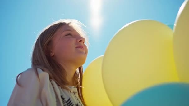 底の角度は屋外の太陽の下で黄色と青の風船で明るい十代の女の子を表示します 雲のない夏の空を背景に笑顔の10代の若者の肖像 — ストック動画