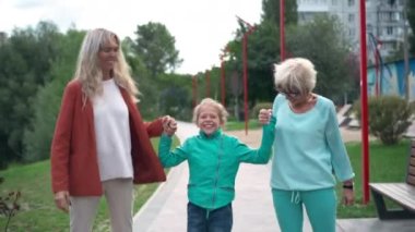 Gülümseyen yetişkin ve son sınıf kadınlarının el ele zıplayarak gülen neşeli kızlar. Neşeli, heyecanlı Kafkasyalı çocuk parkta annesi ve büyükannesiyle eğleniyor.