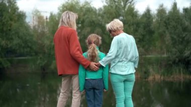 Parktaki göl kıyısında duran anne ve büyükanneye sarılan mutlu kız. Yazın, bulutlu bir yaz gününde, kadınların açık havada olduğundan emin bir beyaz çocuk.
