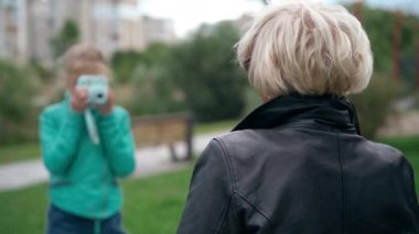 Geçmişe dönüp baktığımızda, gri saçlı yaşlı beyaz bir kadının bulanık kız için fotoğraf çektiğini görüyoruz. Torununu destekleyen pozitif büyükannesinin omzundan vurulmak. Hafta sonları hobi yapmak.