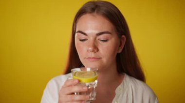 Limonlu su içen genç bir kadın kameraya yavaş çekimde gülümsüyor. Esmer, kendine güvenen, gri gözlü, sarı arka planda sağlıklı içeceklerle poz veren beyaz kadının portresi.