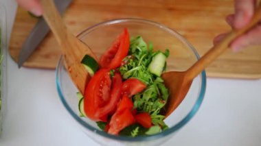 Yakın plan organik sağlıklı salatayı cam kasede yavaş çekimde karıştırmak. Beyaz kadın eller mutfakta sebze karıştırmak için tahta kaşıklar kullanıyor.