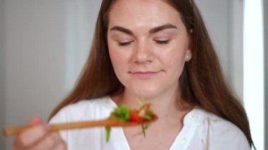 Yakından gülümseyen, salata kokan, gülümseyen, kameralı yiyeceklere bakan bir kadın. Tatmin olmuş beyaz kadın portresi. İçerde lezzetli bir yemekle ağır çekimde poz veriyor.