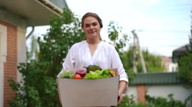 Dışarıda sebze kutusuyla poz veren güzel ve memnun genç bir kadının portresi. Orta ölçekli beyaz kadın elinde organik sağlıklı yiyecek dolu sepetle kameraya bakıyor. Yavaş çekim