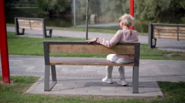 Arka taraftaki dar görüşlü, son sınıf zayıf kadın sağdaki bankta oturuyor. Geniş açılı, kır saçlı, beyaz tenli emekli yaz parkında hafta sonları dışarıda dinleniyor.