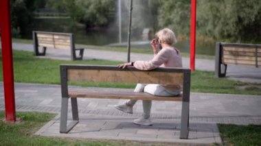 Koronavirüs maskeli yaşlı beyaz kadının açık hava gününde bankta otururken geniş açılı görüntüsü. Gölün kenarındaki parkta dinlenen rahat kadın emekliler.