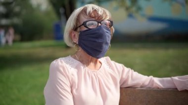 Coronavirus maskeli kıdemli beyaz kadın bankta oturmuş başka tarafa bakıyor ve düşünüyor. Covid-19 salgınında yaşlanan emeklilerin portresi. Yeni normal yaşam tarzı kavramı