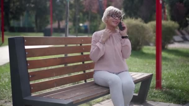 Parktaki Bankta Oturan Gülümseyen Telefonda Konuşan Pozitif Gri Saçlı Kadın — Stok video