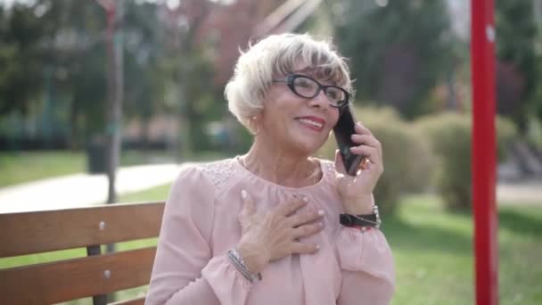 无忧无虑的女性退休人员在户外用电话交谈时触摸着脸的画像 快乐轻松的白种人老年妇女在公园里用智能手机分享流言 — 图库视频影像