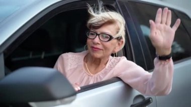 Kıdemli beyaz kadın, şoför koltuğunda oturmuş el sallıyor ve konuşuyor. Otomobilde kendine güveni tam bir emeklinin portresi.