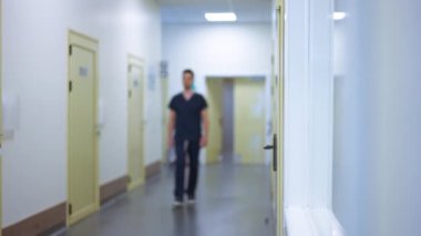 Geniş açılı hastane koridorunda yavaş çekimde yürüyen bulanık bir adam kamerayı geçiyor. Ciddi Orta Doğulu kendine güvenen doktor iş yerinde.