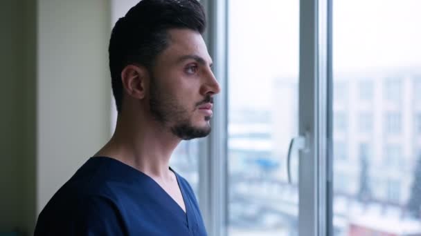 Przemyślany Młody Chirurg Bliskiego Wschodu Patrzący Przez Okno Patrzący Kamerę — Wideo stockowe