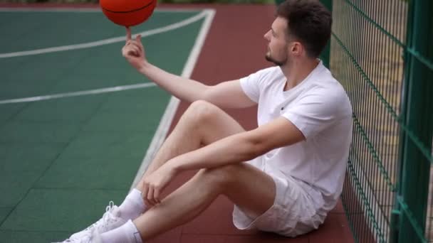指にバスケットボールボールを回転メッシュフェンスに座って若い白人スポーツマンのサイドビューの肖像画 都会の外でハンサムな男の訓練を確信している スローモーション — ストック動画