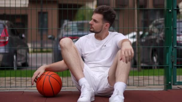 自信に満ちたハンサムな若い男の肖像バスケットボールコートに座って ボールをスローモーションで見て回る フロントビューは都市の屋外でのワークアウトで白人スポーツマンに適合 — ストック動画