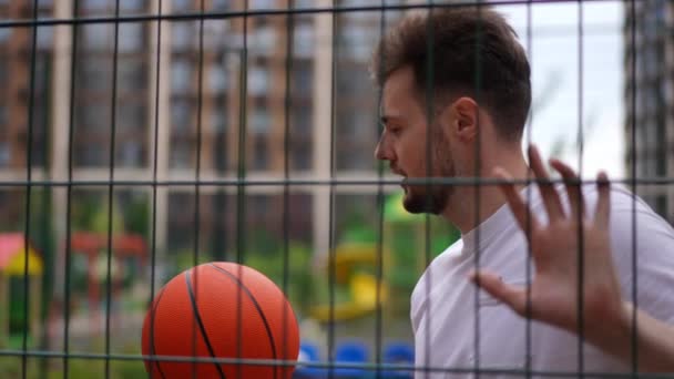 メッシュフェンスに触れる市内のバスケットボールコートにボール立って若いやる気のあるスポーツマンのサイドビューの肖像画 設備屋外で白人選手を確信してください スローモーション — ストック動画