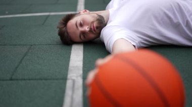 Yeşil basketbol sahasında yatan yakışıklı genç adam topa dokunup sırtını dönüyor. Beyaz tenli sporcunun şehir dışında antrenman yapması çok düşünceli bir hareket. Spor ve motivasyon kavramı