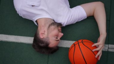 Top view sporcusu yeşil spor sahasında turuncu basketbol topuyla yatarken yavaş çekimde canlı kamera dönüyor. Dışarıdaki kendine güvenen beyaz yakışıklı adamın portresi.