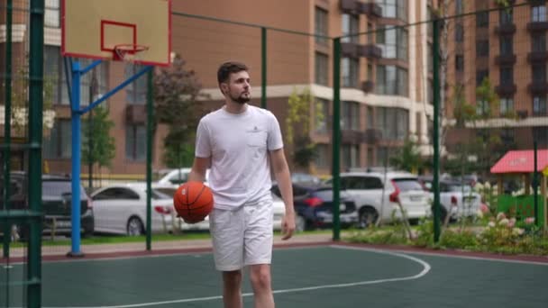 バスケットボールボールを屋外に残して歩く自信のあるスポーツマンのフロントビューの肖像画 屋外裁判所で都会を散歩している白人の若者に合う 動機とスポーツの概念 — ストック動画