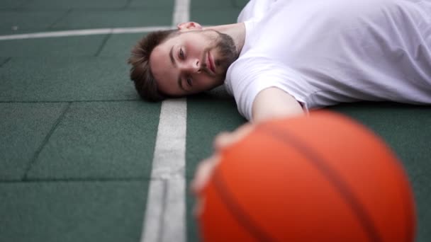 緑のバスケットボールコートに寝そべっているハンサムな若い男がボールに触れて背を向けている 市内の屋外でトレーニングを中断している白人のスポーツマンにふさわしいと考えられます スポーツとやる気のコンセプト — ストック動画