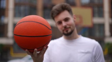 Yakın plan basketbol topu ve erkek eli arka planda bulanık gülümseyen bir sporcuyla kamerayı işaret ediyor. Kafkas formundaki bir adam açık havada reklam oyunu oynuyor.