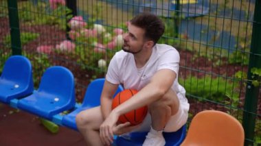 Beyaz tenli sporcunun, basketbol topuyla dışarıda otururken kameraya bakarken gülümsediği yüksek açılı zoom. Genç formda adam spor malzemeleriyle ağır çekimde poz veriyor.