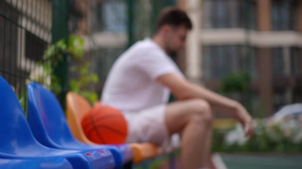 バスケットボールのボールに座ってぼやけた白人のスポーツマンとスポーツの地面に椅子の側面の角度ビューの行 認識できないフィット若いです男オンブレークトレーニングで街屋外 — ストック動画
