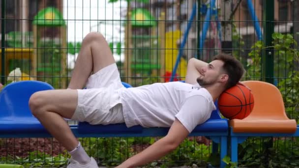 サイドビューワイドショット笑顔幸せなスポーツマンの頭の下にバスケットボールボールと椅子に横たわっている 自信を持って訓練に休憩を考えて離れて見て白人男性に適合します 仕事とモチベーション — ストック動画