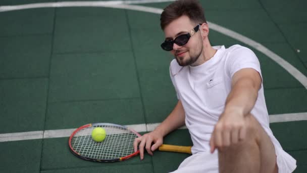 遅い動きでサングラスを脱いでテニスコートに座って笑顔カメラを見てハンサムな若いスポーツマン ラケットとボールで屋外でポーズの満足成功した選手の肖像 — ストック動画