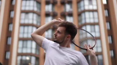Kafasının arkasında tenis raketi olan kendine güveni tam genç sporcu açık şehir binasının arka planında duruyor. Etrafı kolaçan eden yakışıklı beyaz oyuncunun portresi.
