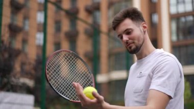 Genç, gülümseyen, zinde, sağda duran sporcu elinde tenis topuna bakıyor ve başka tarafa bakıyor. Şehir merkezindeki tenis kortunda motive olmuş beyaz bir adamın portresi.