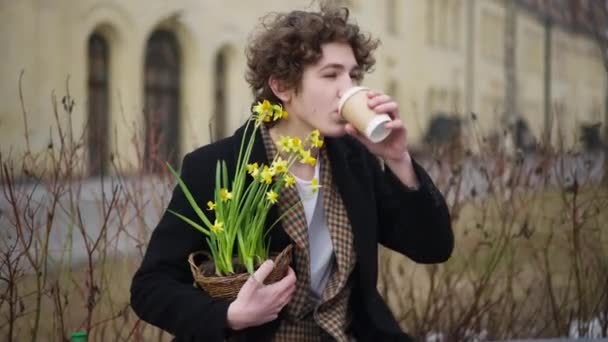 中ショット笑顔男コーヒーを飲んでポットに黄色の花と市内のベンチに座っている 自信満足の肖像画幸せな白人エレガントな男屋外でドリンクと植物 — ストック動画