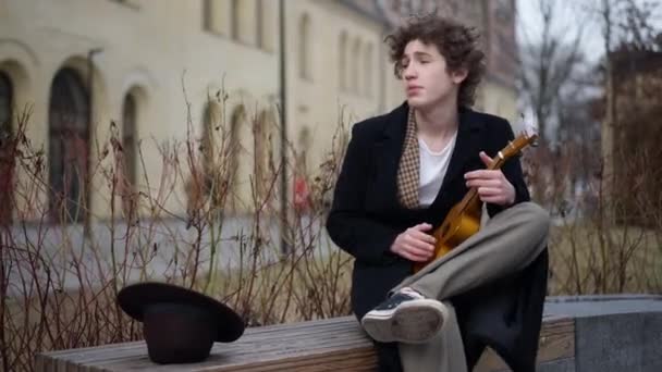 都市部のベンチに座って歌うウクレレを演奏若い才能の男の肖像画 風の強い曇りの日に屋外でレジャーを楽しむハンサムな白人歌手のミュージシャン — ストック動画