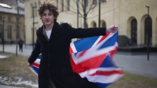 ミディアムショット喜びの若い男都市の街にイギリスの旗を掲揚 リラックスした陽気な白人男の肖像写真を見ますカメラ笑顔立って屋外でスティック舌アウト — ストック動画