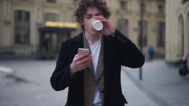 フロントビューは スマートフォンアプリのコーヒーを飲むの街の通りのメッセージングをオンラインで歩く若い男が集中した ハンサムな白人エレガントな男散歩屋外で朝スクロールソーシャルメディア — ストック動画