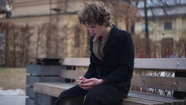 問題を抱えた若いハンサムな男のベンチに街の通りのメッセージをオンラインで離れて見て考えて座っている 不確定な躊躇の肖像 電話アプリでインターネットサーフィン白人エレガントな男屋外 — ストック動画