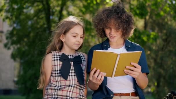 Щасливий Кавказький Хлопчик Читає Вголос Книжку Посміхається Дівчина Сидить Лавці — стокове відео